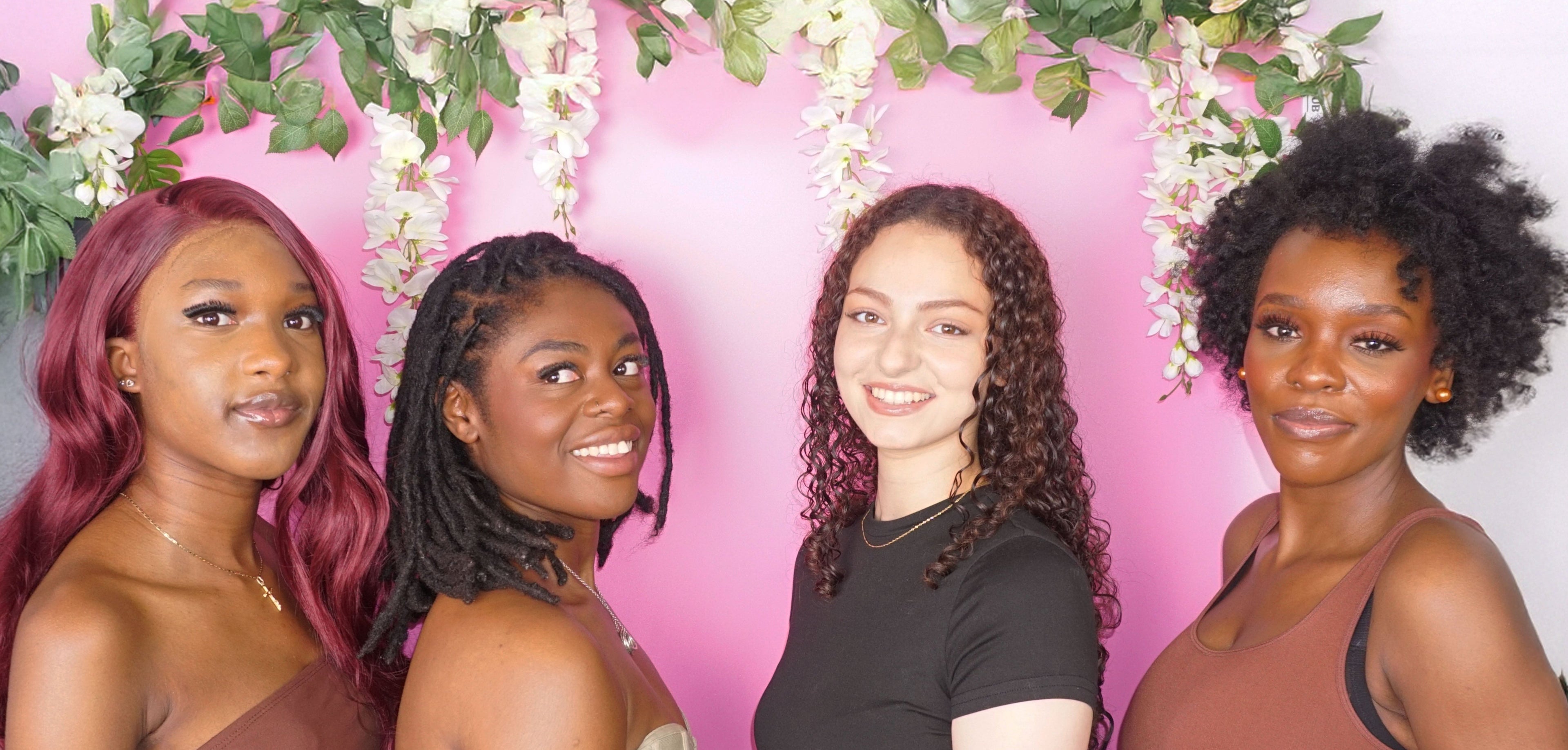 Four women with diverse hairstyles standing in front of a pink background with floral decorations.