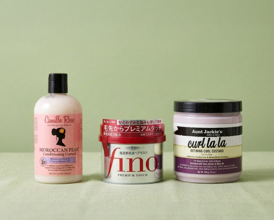 Three hair care products arranged side by side on a light green surface against a matching green background: a pink bottle labeled Curly Girl Moroccan Hair, a white tub labeled Vino, and a white jar labeled Aunt Jockie's Curl & Co.