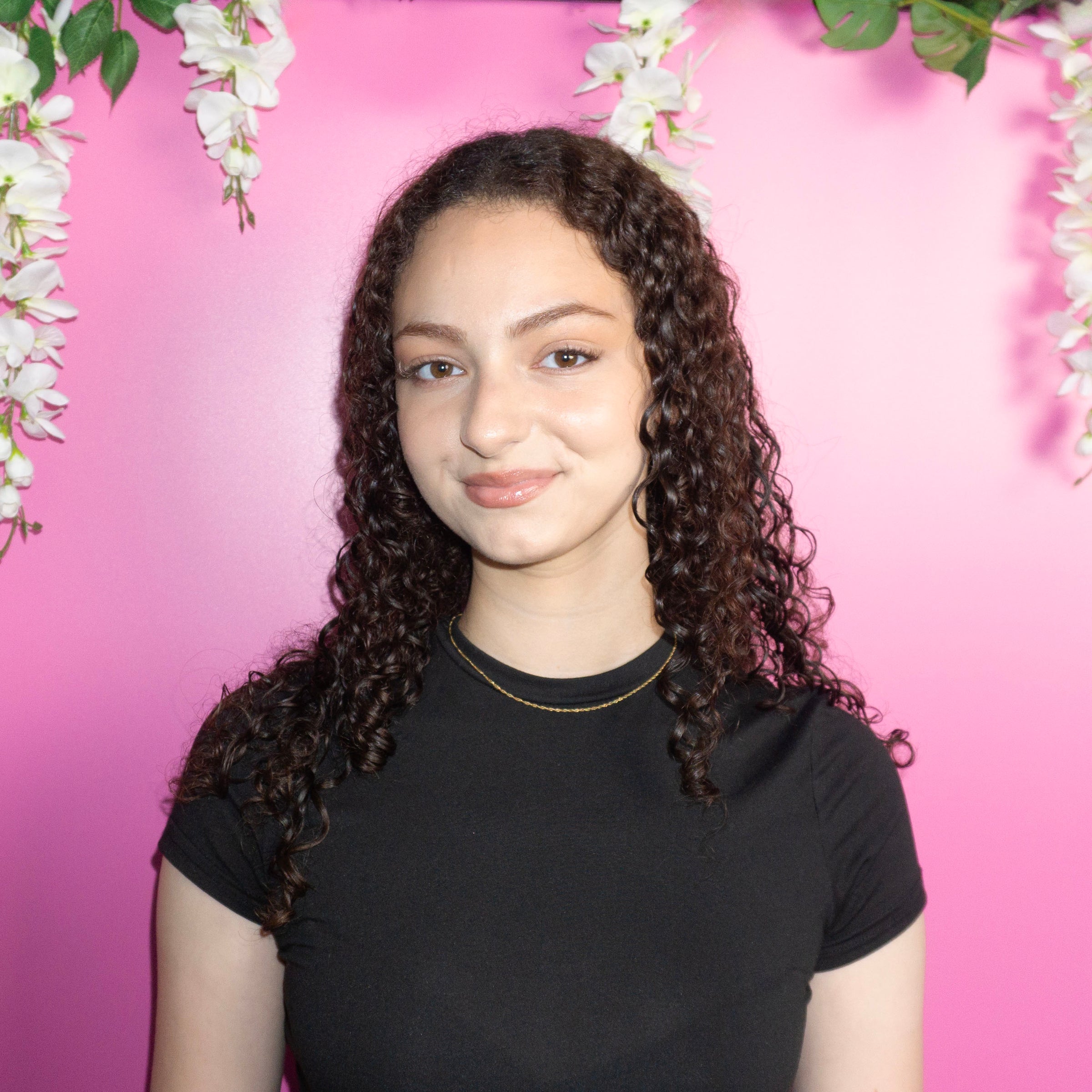 Person with curly hair wearing a black shirt against a pink background with floral decorations